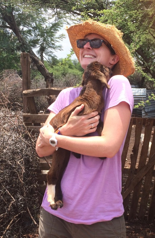 Woman with hat and sunglasses holding a brown baby goat in her arms and smiling.